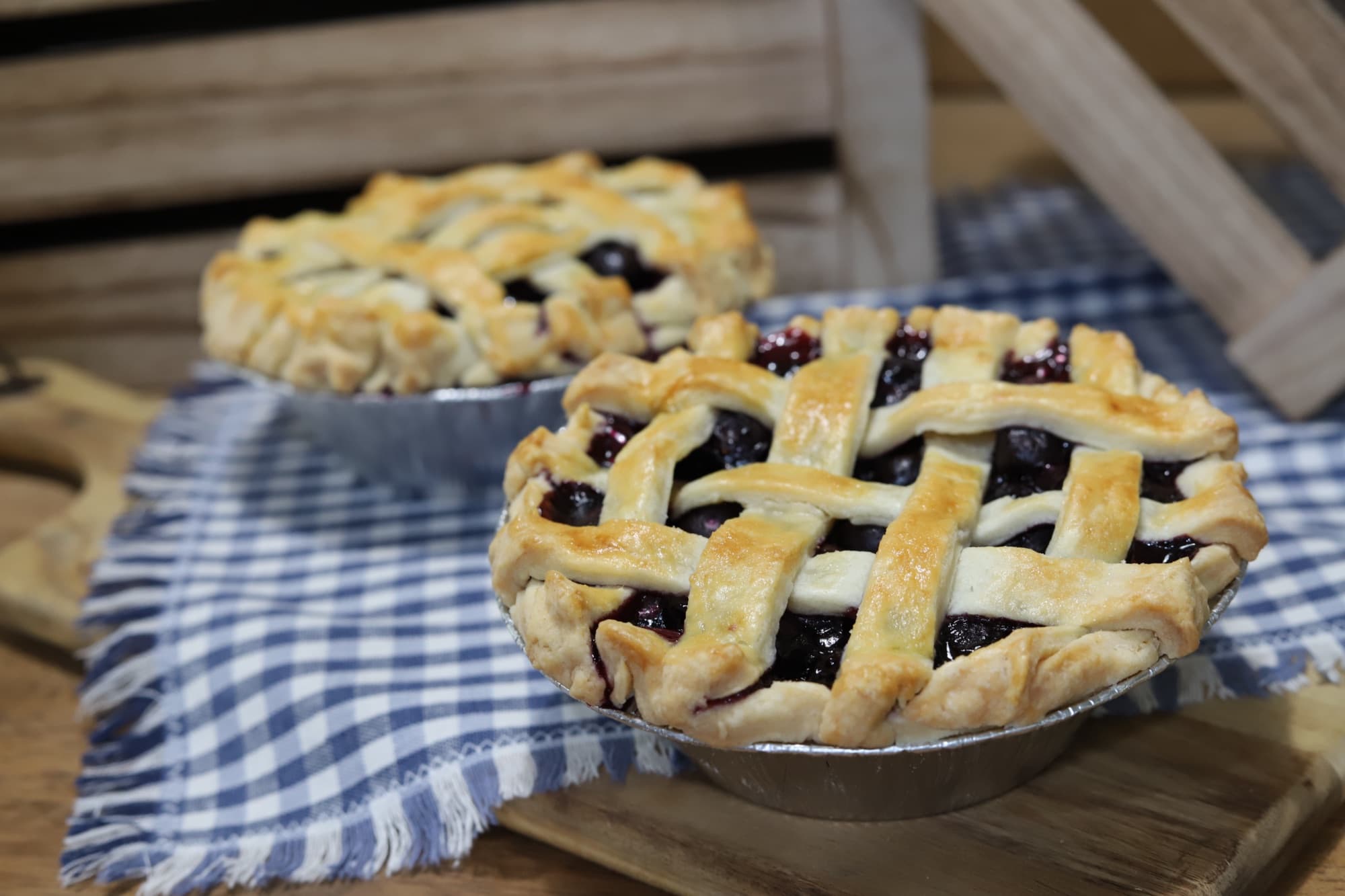 Two lattice-topped blueberry pies cooling on a gingham cloth